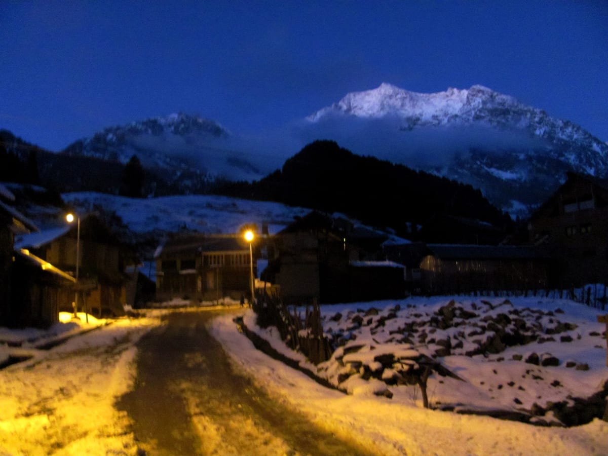A snowy village road at night in Aru Valley, warm lights glowing beneath moonlit peaks